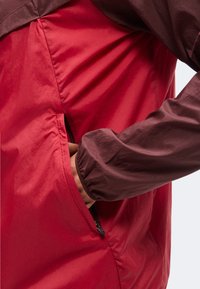 Une personne portant une veste rouge avec une poche zippée, la main partiellement dans la poche, la manche visible en tissu marron foncé.