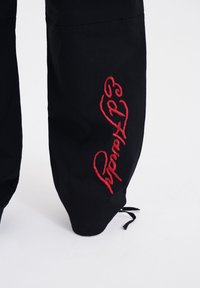 Czarne spodnie z czerwonym haftowanym napisem "Ed Hardy" wzdłuż nogawki, o luźnym kroju i regulowanym sznurkiem u dołu.