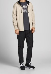 Felpa beige con accenti neri, t-shirt grafica grigia sotto, pantaloni cargo neri e sneakers nere con lacci bianchi.