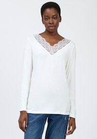 Hvid langærmet top med V-hals, der har en delikat blondekant. Stoffet er blødt, med en tætsiddende silhuet og subtile syninger.
