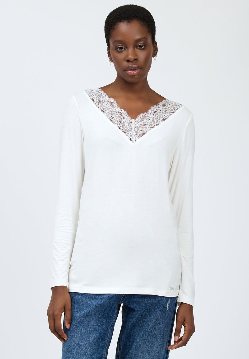 Hvid langærmet top med V-hals, der har en delikat blondekant. Stoffet er blødt, med en tætsiddende silhuet og subtile syninger.