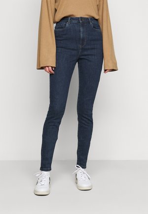 Osoba ubrana w ciemnoniebieskie obcisłe dżinsy, luźną beżową bluzkę z długim rękawem i białe trampki, stojąca na jednolitym tle.