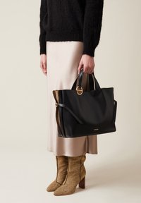 Schwarze Leder-Tote mit einem strukturierten braunen Akzent, runden Griffen und einem goldenen Hardware-Detail, gehalten neben einem Satinrock und gemusterten Stiefeln.