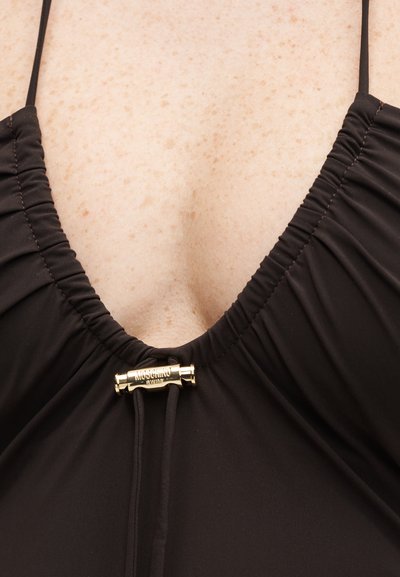 Κοντινό πλάνο μαύρου μαγιό με ρίγες στη λαιμόκοψη, λεπτές τιράντες και χρυσή διακοσμητική αγκράφα με χαραγμένο "Moschino Swim."