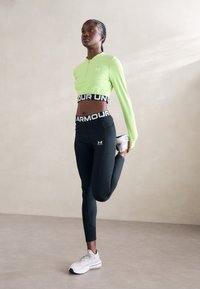 Világos zöld, hosszú ujjú crop top cipzárral, fekete, magas derekú leggings fehér márkázással, és szürke sportcipő. Nyújtó póz.