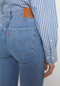 Vaqueros de mezclilla azul claro con cintura alta, bolsillos traseros en forma de corazón y una etiqueta de cuero marrón. Presentan un sutil desgaste y detalles de costura.