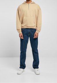 Persona con felpa beige con cappuccio e tasca frontale, jeans blu e sneakers bianche, in piedi contro uno sfondo grigio neutro.