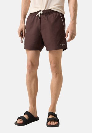 Mand iført brune badeshorts med hvid snor, holder solbriller og sorte sandaler, står foran en ensfarvet baggrund.