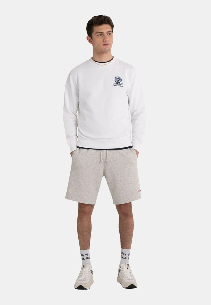 Sweat à capuche blanc avec logo sur la poitrine, associé à un short gris. Le modèle porte des baskets blanches et des chaussettes à motifs. Look de sport simple et décontracté.