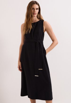Vrouw met lang bruin haar die een mouwloze zwarte midi-jurk draagt met een gestrikte tailleband, staand tegen een effen lichte achtergrond.