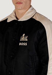 Giacca in raso nero con colletto crema. Presenta i loghi "SUPER BOWL" e "BOSS" in crema. Include bottoni a pressione e tasche laterali.