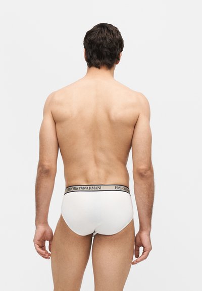 Άνδρας στέκεται με την πλάτη, φοράει λευκά εσώρουχα Emporio Armani με μαύρη ζώνη με λογότυπο πάνω σε απλό λευκό φόντο.