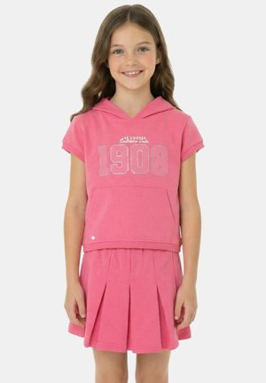 Fille souriante aux longs cheveux bruns portant un haut à capuche rose à manches courtes avec "1908" et une jupe plissée rose assortie.