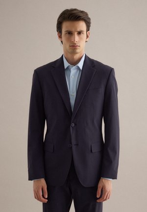 Jonge man in een donkerblauw colbert en lichtblauw overhemd, staand met ontspannen armen tegen een effen beige achtergrond.