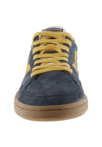 Zapatillas de ante azul marino con cordones y acentos amarillos, etiqueta en la lengüeta blanca y suela de goma texturizada. Presenta perforaciones en la parte superior.