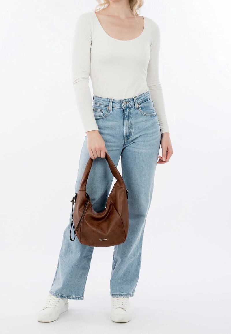 Femme portant un haut blanc à manches longues, un jean taille haute bleu clair, des baskets blanches, tenant un sac à main en cuir marron par ses anses.