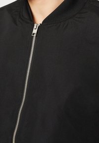 Veste bomber noire avec une fermeture éclair métallique argentée et un col côtelé, montrée fermée sur le cou et les épaules d'une personne.