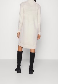 Abito lavorato a maglia beige con un alto colletto, texture a coste sulle spalle e maniche lunghe. Abbinato a stivali neri al ginocchio.