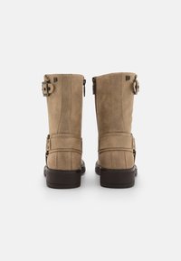 Bottes en suede tan jusqu'au genou avec un talon plat marron, dotées de deux boucles ajustables et d'une fermeture éclair sur le côté pour un accès facile.