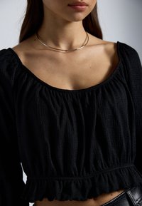 Svart teksturert bluse med samlet halslinning og volangkant, med lange ermer og avslappet passform. En enkel metallisk choker-halskjede bæres.