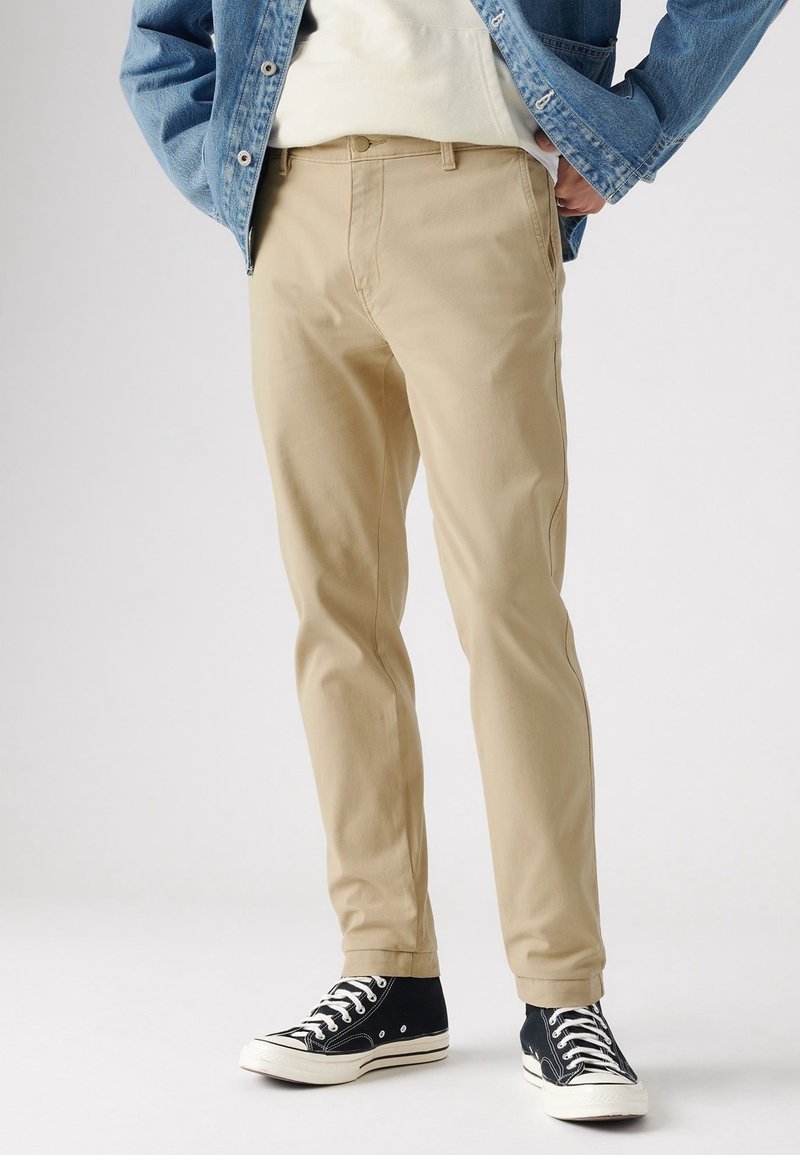 Homme portant un pantalon beige à coupe droite, des baskets montantes noires, un sweat à capuche blanc et une veste en jean, debout les mains dans les poches.