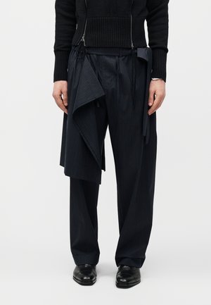 Άτομο που φοράει μαύρο παντελόνι με λευκές ρίγες, φαρδιά μπατζάκια, με ασύμμετρη στρωμένη ύφανση πάνω από το ένα πόδι και μαύρα επίσημα παπούτσια.