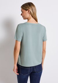 Krátk�ý rukáv, světle zelený top vyrobený z měkké tkaniny, kulatý výstřih, pohodlný střih, hladká textura, s rovný lem. Nosený přes tmavé džíny.