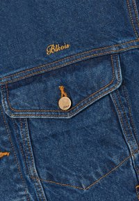 Poche de veste en denim avec surpiqûres orange, un bouton portant l'inscription "Bbkvis" et le texte "Bbkvis" brodé au-dessus du rabat de la poche.