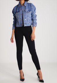 Veste en denim bleu clair avec manches longues, design ajusté, deux poches poitrine, associée à un legging noir et des chaussures à talons.