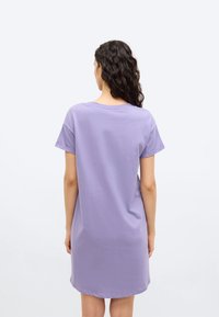 Γυναίκα με μακριά σγουρά μαλλιά, φορώντας ένα λιλά φόρεμα με κοντά μανίκια στο μήκος του γονάτου, γυρισμένη από πίσω σε ένα απλό λευκό φόντο.