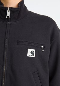 Giacca nera con zip realizzata in tessuto spesso, dotata di un colletto alto, una piccola tasca frontale con zip e un logo ricamato sul petto.