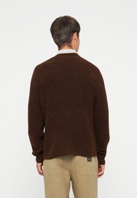Brown knitted sweater with a textured pattern, long sleeves, and a crew neck, paired with light khaki pants featuring a pocket label.
