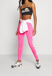 Czarny krótki top z białym logo, połączony z neonowymi różowymi legginsami z białym napisem. Biała koszula związana w talii i masywne trampki.