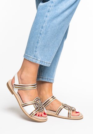 White sandals with gold and brown accents, featuring a decorative knot design. Textured sole, adjustable straps, and open-toe style.