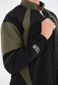 Svart og olivengrønn jakke med høyt krage, elastiske mansjetter, glidelås og et merke på ermet. Lettvektig stoff.