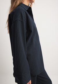 Camicia blu navy con bottoni, caratterizzata da sottili righe verticali bianche, colletto, maniche lunghe e una vestibilità comoda.