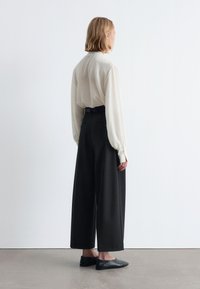 White, long-sleeve shirt with a high neckline paired with wide-leg black trousers, featuring a belted waist and flat black shoes.