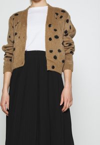 Brun oversized cardigan i tekstureret strik, med sorte prikker og sorte knapper, parret med en hvid skjorte og en sort plisseret nederdel.