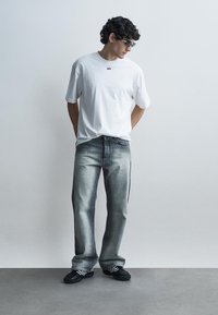 Fiatal férfi göndör hajjal, napszemüveget viselve, egy fehér, túlméretezett pólót, bő, kifakult farmert és fekete sportcipőt visel, egyszerű háttér előtt áll.