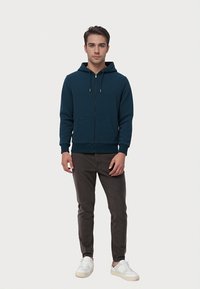 Marineblå zip-up hættetrøje med frontlommer, snoretræk i hætten, ribbede manchetter og talje; parret med mørkegrå bukser og hvide sneakers.