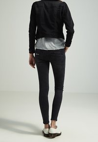 Chaqueta de denim negra llevada sobre una camisa gris, combinada con jeans oscuros ajustados y zapatos blancos. Bolsillos traseros visibles en los jeans.
