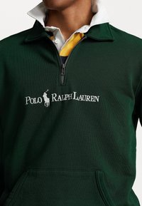 Pullover verde con zip al collo, caratterizzato da un logo ricamato bianco "Polo Ralph Lauren". Il colletto è stratificato con una maglietta bianca sottostante.
