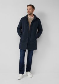 Navyblauwe, middellange jas met een spread collar, knoopsluiting aan de voorkant en zijzakken. Draagt over een beige gebreide trui en blauwe spijkerbroek.