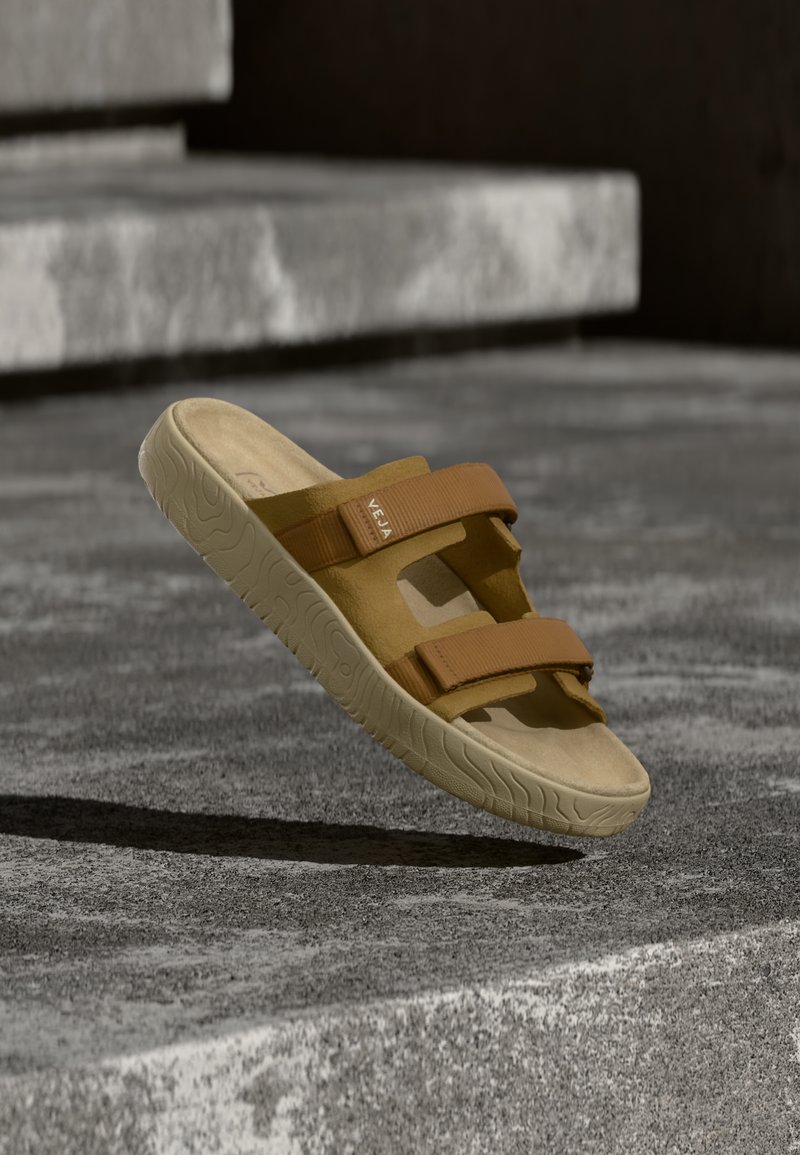 Καφέ σανδάλια με δύο ρυθμιζόμενους ιμάντες και υφή στο πέλμα. Κατασκευασμένα από μαλακό υλικό, με κοψίματα και μινιμαλιστικό σχεδιασμό.