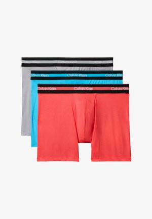 Muskelöser Mann steht barfuß vor grauem Hintergrund, trägt hellgraue Boxer-Shorts mit schwarzem Bund, Arme vor der Brust verschränkt.