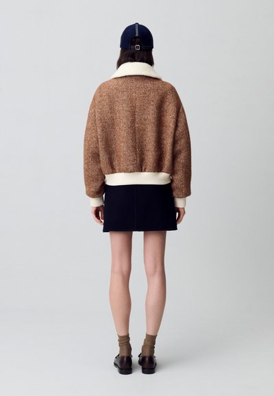 Suéter de textura en espiga marrón con cuello blanco; combinado con una falda azul marino, calcetines marrones y zapatos oscuros, visto desde atrás.