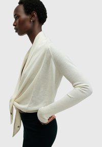 Cárdigan ligero color crema con un frente drapeado, mangas largas, puños acanalados y abertura en la parte trasera. Diseño sencillo con detalles de botones.