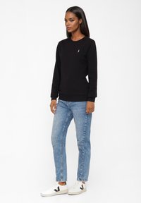 Zwarte sweatshirt met lange mouwen, geribde manchetten en zoom. De sweatshirt is gecombineerd met lichtblauwe jeans en witte sneakers met zwarte accenten.