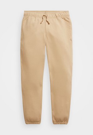 Beige afslappede joggingbukser med elastisk taljebånd, snøre, sidelommer og elastiske manchetter, lagt fladt på en hvid baggrund.