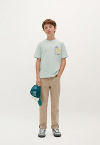 Lichtgroene t-shirt met gele schedelafbeelding, beige broek, grijze en blauwe schoenen, en een turquoise mesh pet in de hand.